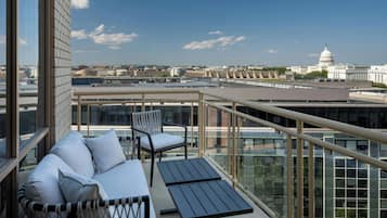 A balcony with a sofa and chairs, overlooking a cityscape with a prominent white building.