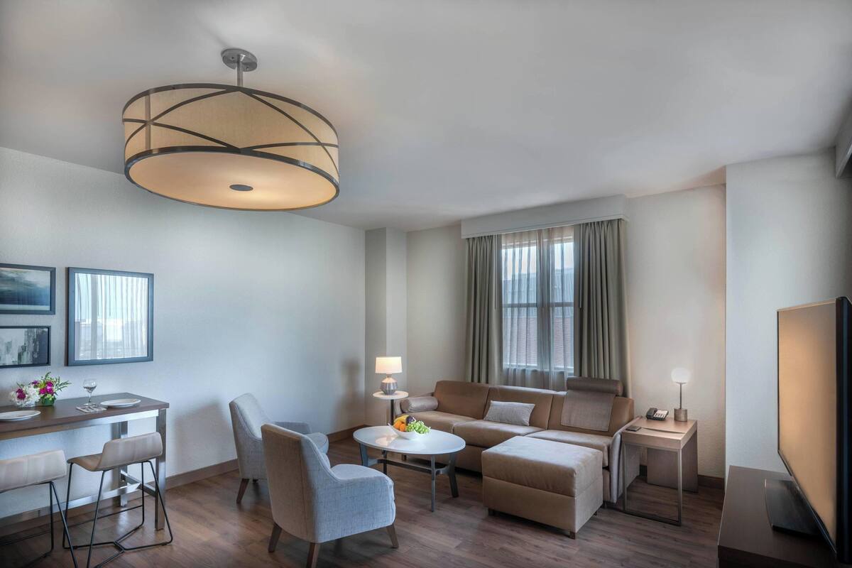 Una habitación de hotel moderna con zona de comedor, sofá, televisor y un amplio ventanal con cortinas.