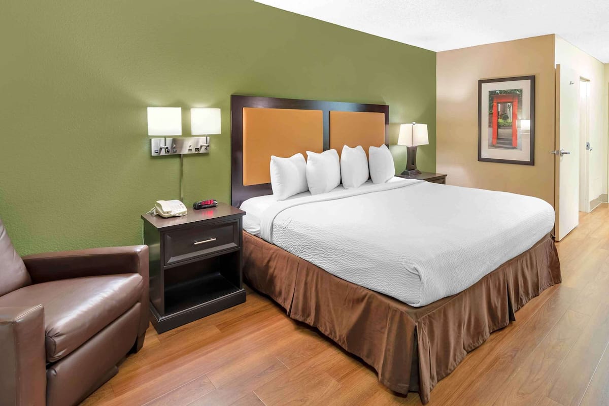 A hotel room with a large bed, a nightstand with a lamp, a chair, and a framed picture on the wall.
