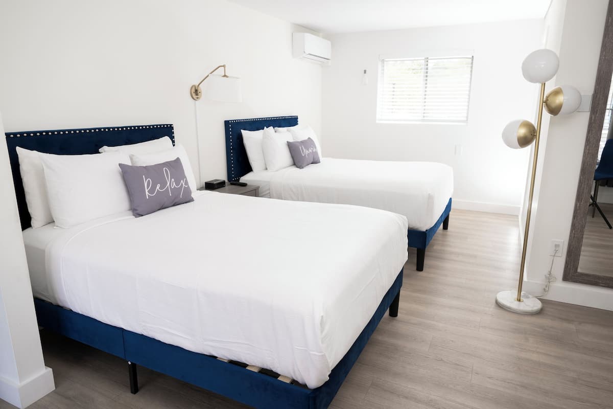 A hotel room with two beds, each with a blue headboard and white bedding. There is a floor lamp with a gold base and white lampshade.