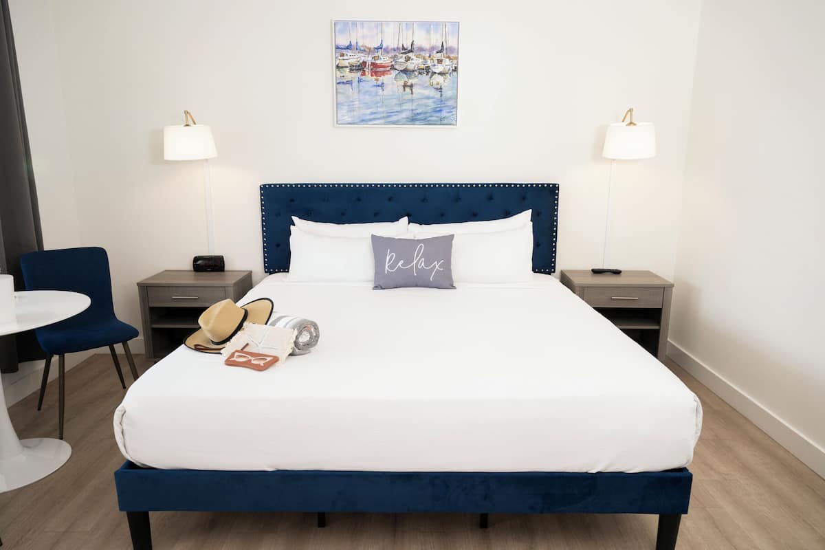 A neatly made bed with a blue headboard, two bedside tables with lamps, a chair, and a painting of boats on the wall.