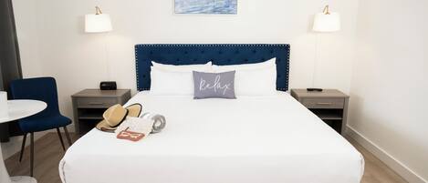 A neatly made bed with a blue headboard, two bedside tables with lamps, a chair, and a painting of boats on the wall.