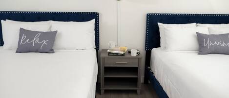 Une chambre d’hôtel avec deux lits, chacun recouvert d’une couette blanche et doté d’oreillers, une table de chevet avec une lampe et une petite étagère avec des livres et une tasse.