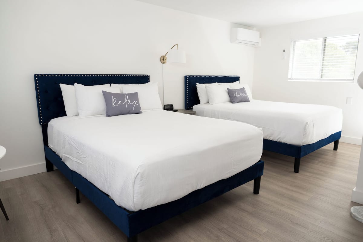 Two beds with dark blue headboards and white bedding, a bedside table, a lamp, and a window with blinds.
