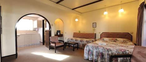 A hotel room with two beds, a sitting area, a small table, and a kitchenette visible through an archway.