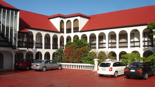 Un edificio de paredes blancas y techo rojo, con múltiples ventanas de arco y balcones. Varios coches estacionados frente a él.