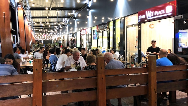 A bustling indoor dining area with wooden benches and tables, ceiling fans, and string lights.