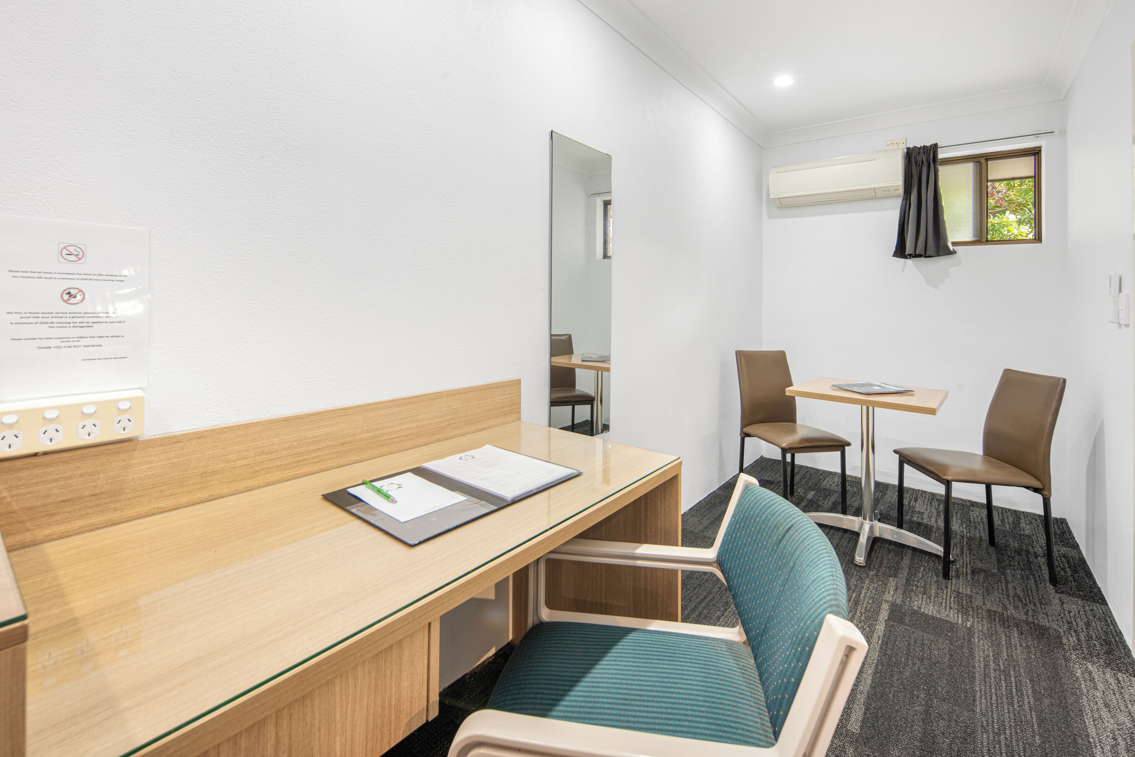 A hotel room with a wooden desk, a chair, a small table, and a window with curtains.