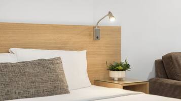 Una cama bien hecha con almohadas blancas y con textura, una mesita de noche con una planta y un sillón tapizado en tono café.