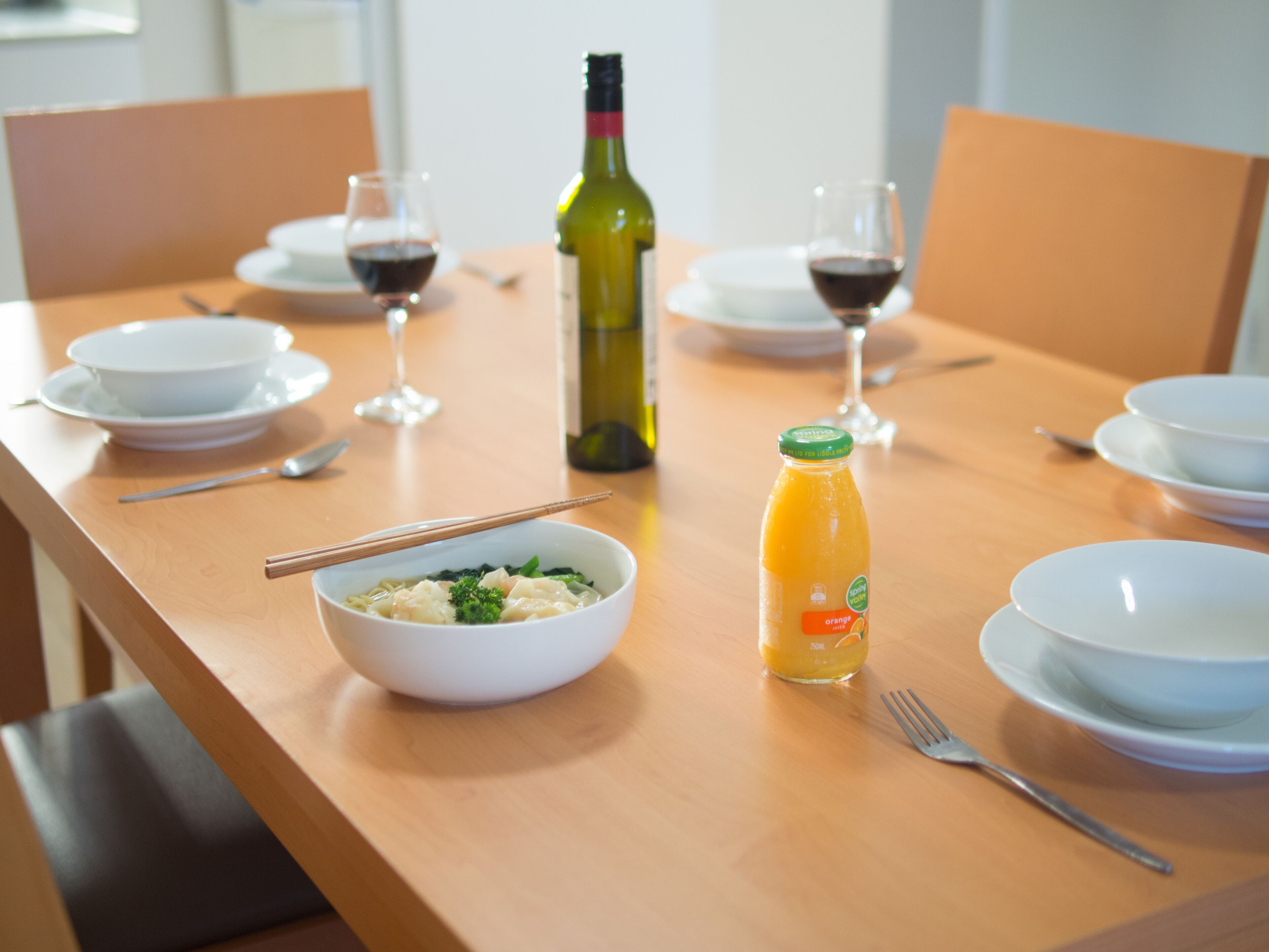 Una mesa de comedor con cuencos, palillos, una botella de jugo de naranja y copas de vino.