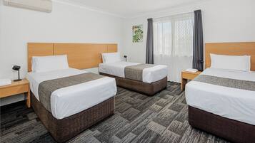 A hotel room with three single beds, a bedside table with a lamp, a wall-mounted air conditioner, and a window with curtains.