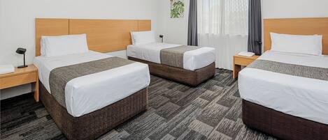 Habitación de hotel con tres camas individuales, una mesita de noche con lámpara, aire acondicionado instalado en la pared y una ventana con cortinas.
