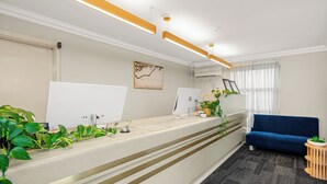 A reception desk with a flat-screen monitor, a desktop computer, and a telephone. There are plants on the desk, a blue sofa, and a window with curtains.