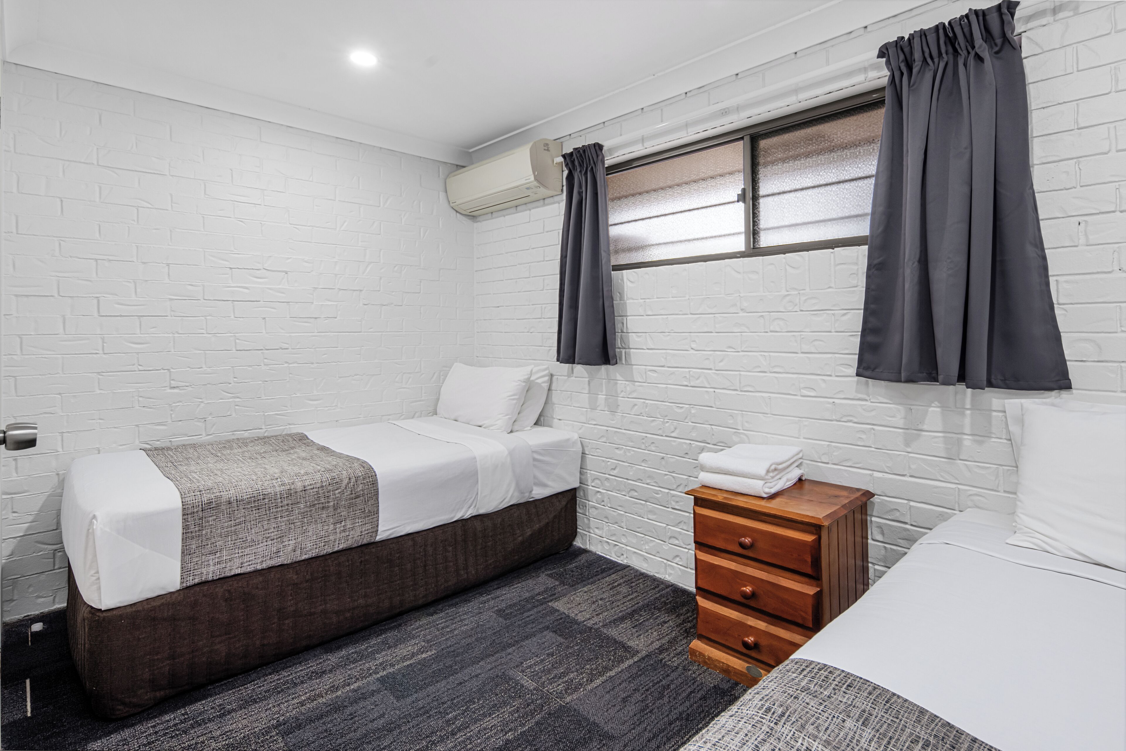A bedroom with a bed, a bedside table, a dresser, a window with curtains, and a brick-patterned wall.