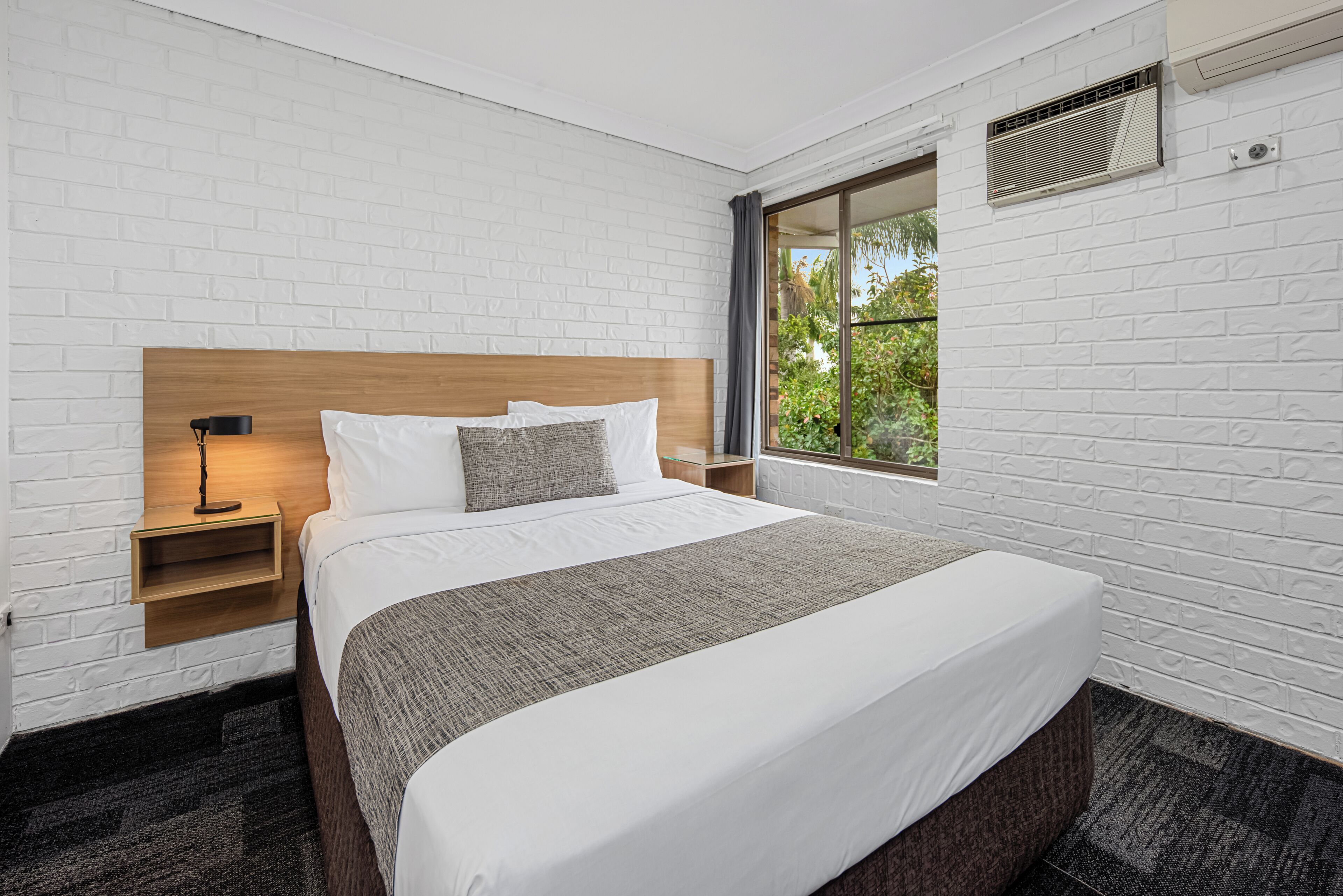 A hotel room with a large bed, a nightstand, a lamp, and a window with a view of greenery.