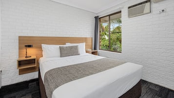 A hotel room with a large bed, a nightstand, a lamp, and a window with a view of greenery.