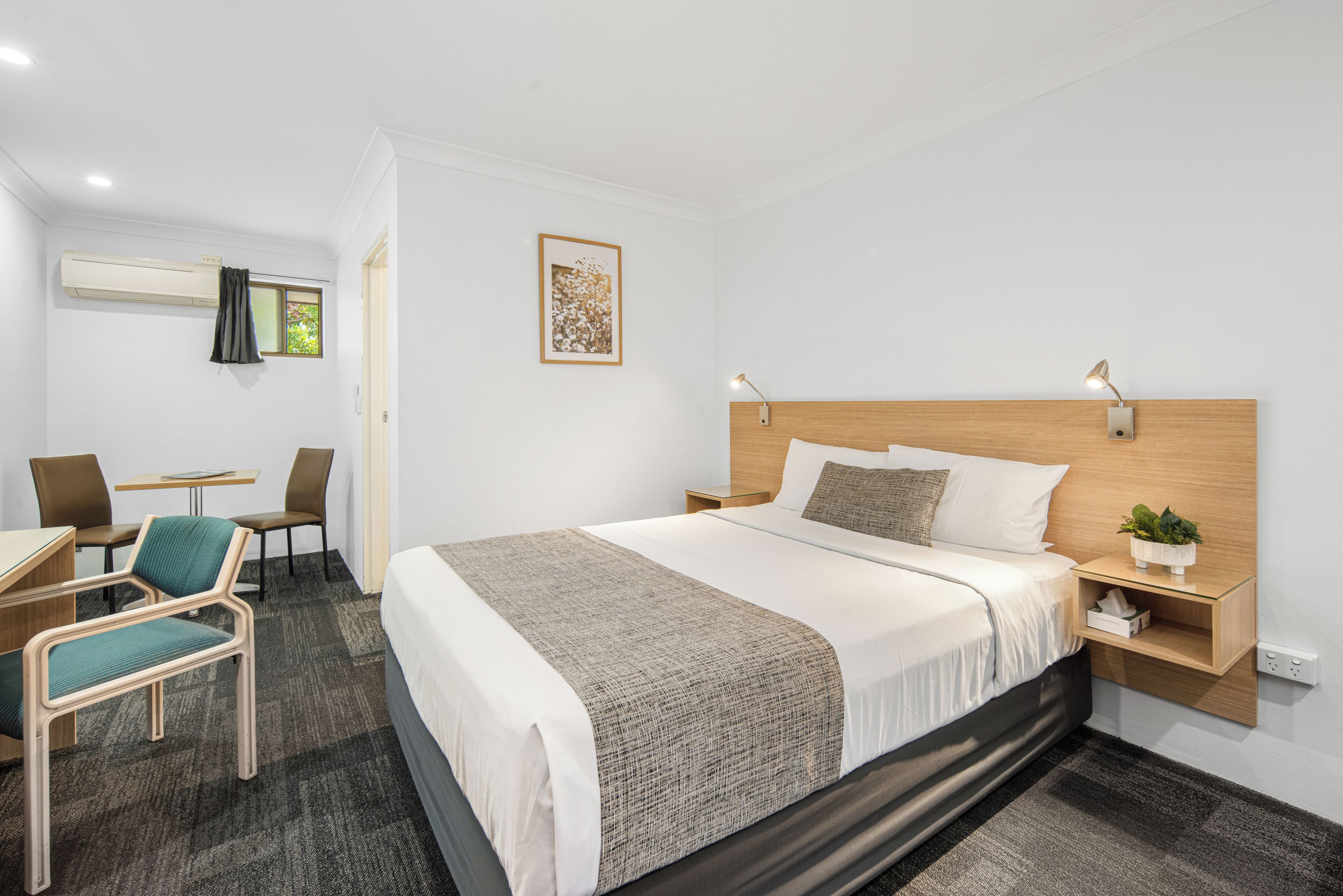 A hotel room with a bed, bedside tables, a desk with a chair, an air conditioner, and a window with curtains.