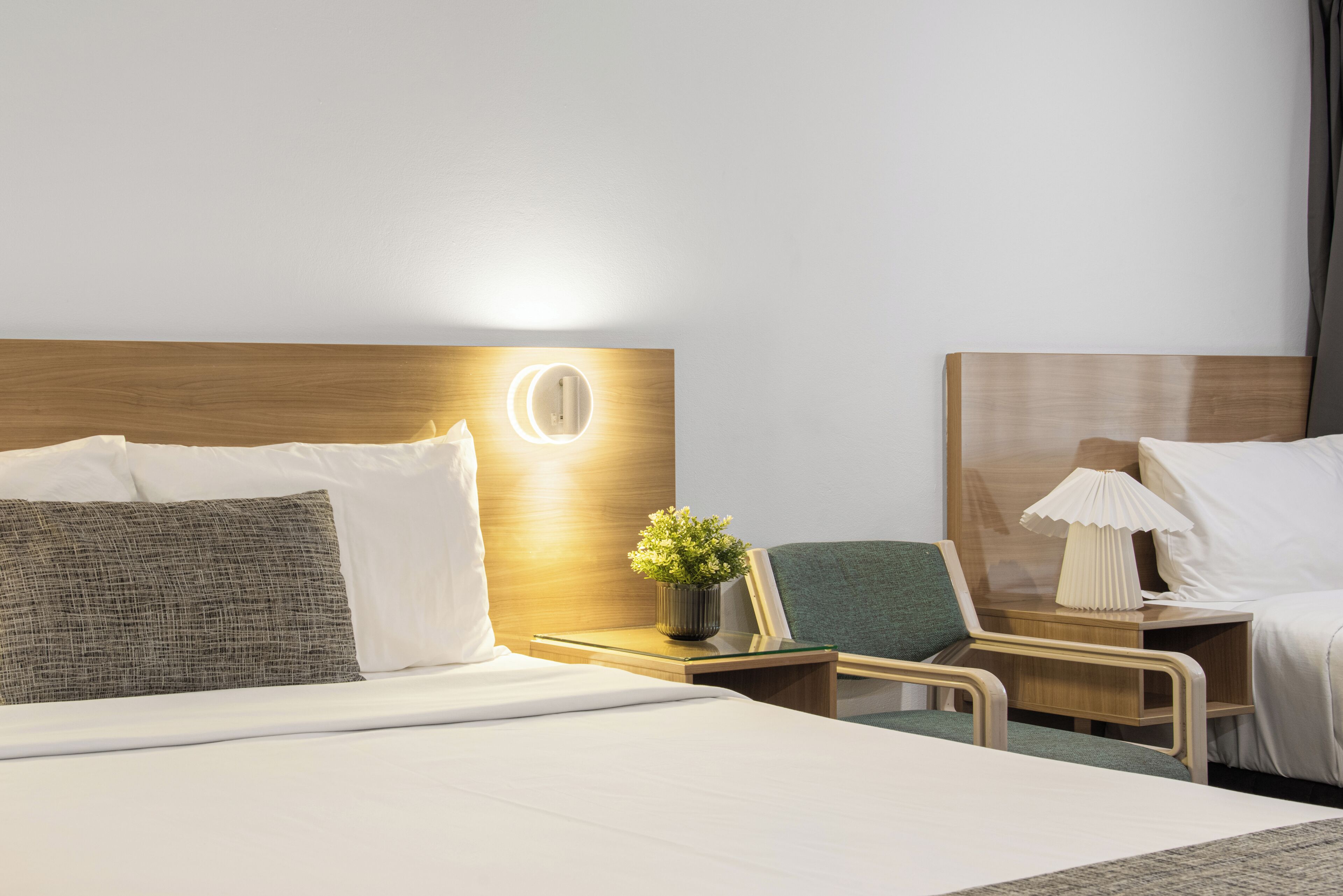 Habitación de hotel con una cama grande, una mesita de noche con lámpara, una silla pequeña y una mesita auxiliar.