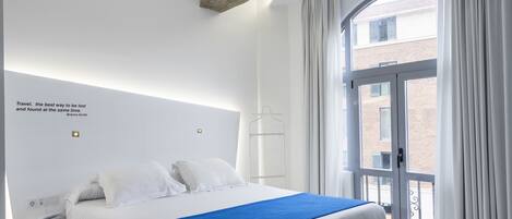 A modern hotel room with a large bed, a view of the city through a large window, and a quote on the wall.