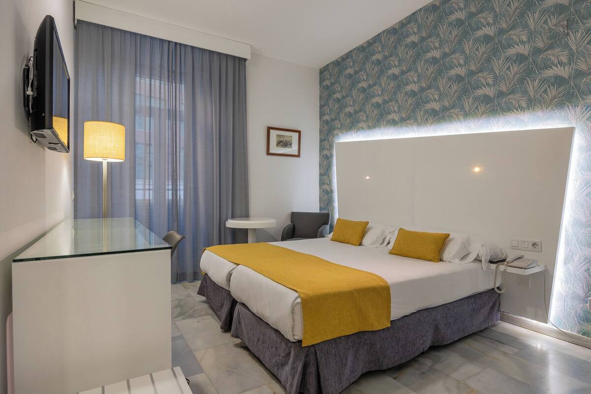 A hotel room with a large bed, a bedside table, a desk, a chair, a mirror, a lamp, a telephone, and a picture on the wall.