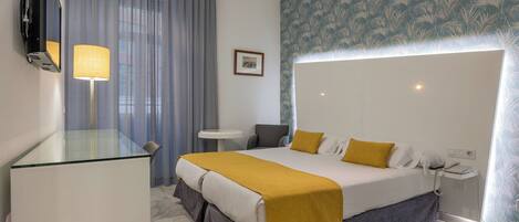 A hotel room with a large bed, a bedside table, a desk, a chair, a mirror, a lamp, a telephone, and a picture on the wall.