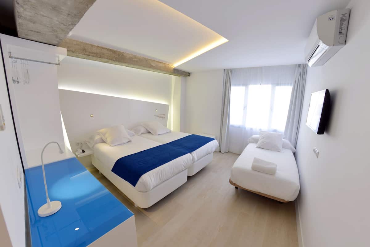 A modern hotel room with a large bed, a smaller bed, a desk with a lamp, and a flat-screen TV mounted on the wall.