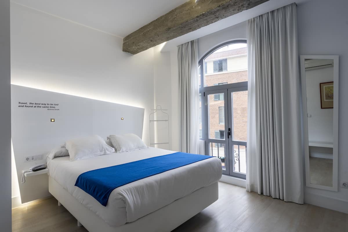 A modern hotel room with a large bed, a blue throw blanket, a white nightstand, a wall-mounted light, and a large window with curtains.