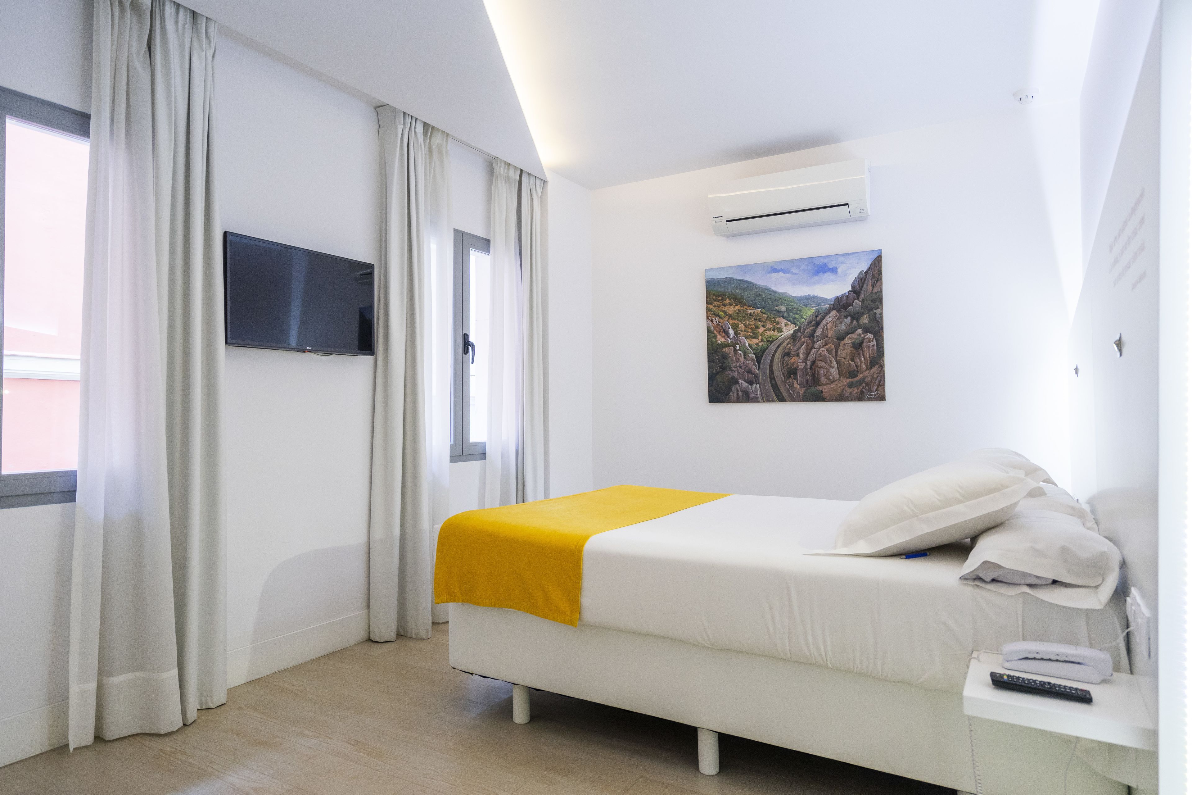 A hotel room with a bed, a TV mounted on the wall, an air conditioning unit, and a painting of a landscape.