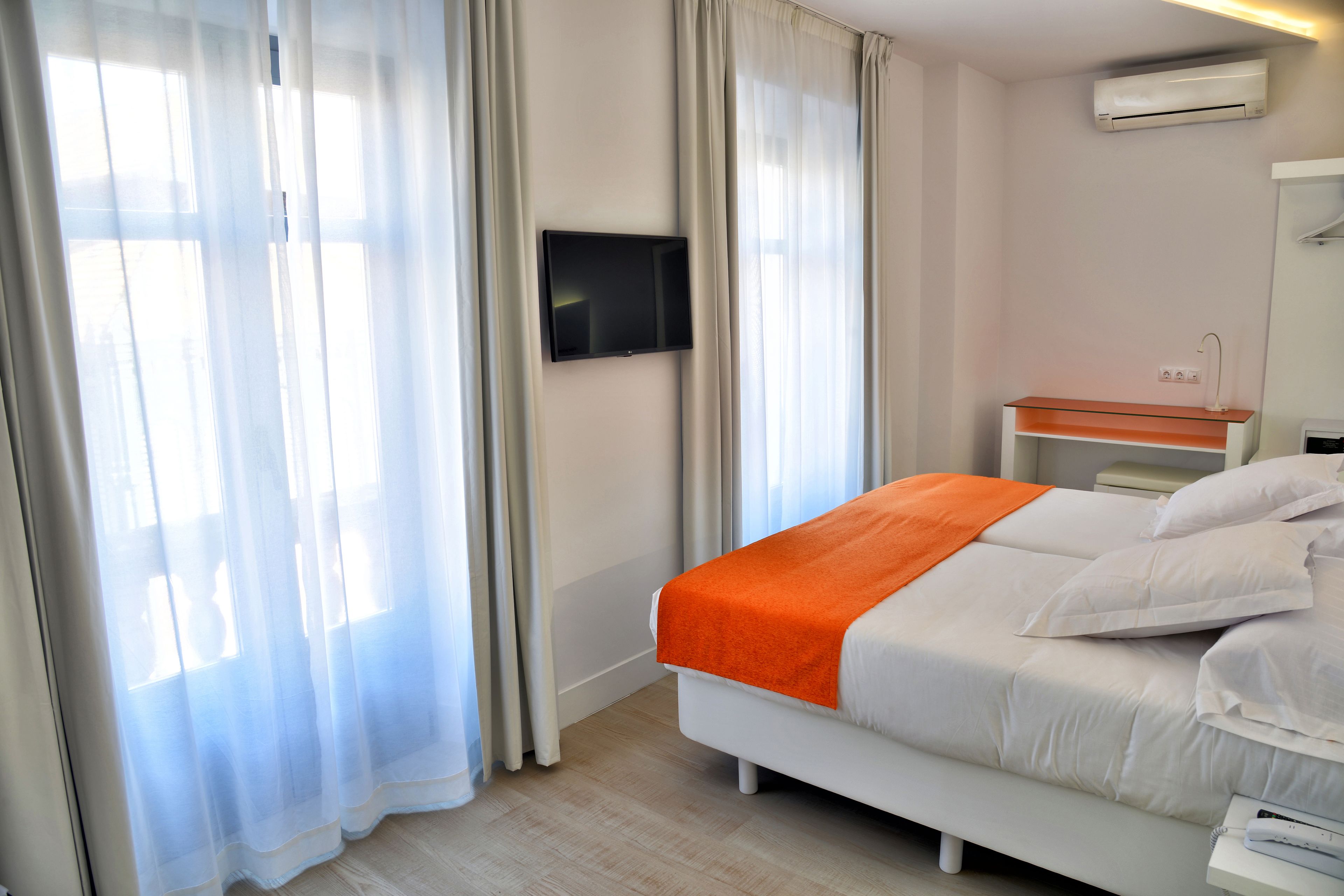 A hotel room with a bed, a TV mounted on the wall, a bedside table with a lamp, and an air conditioning unit.
