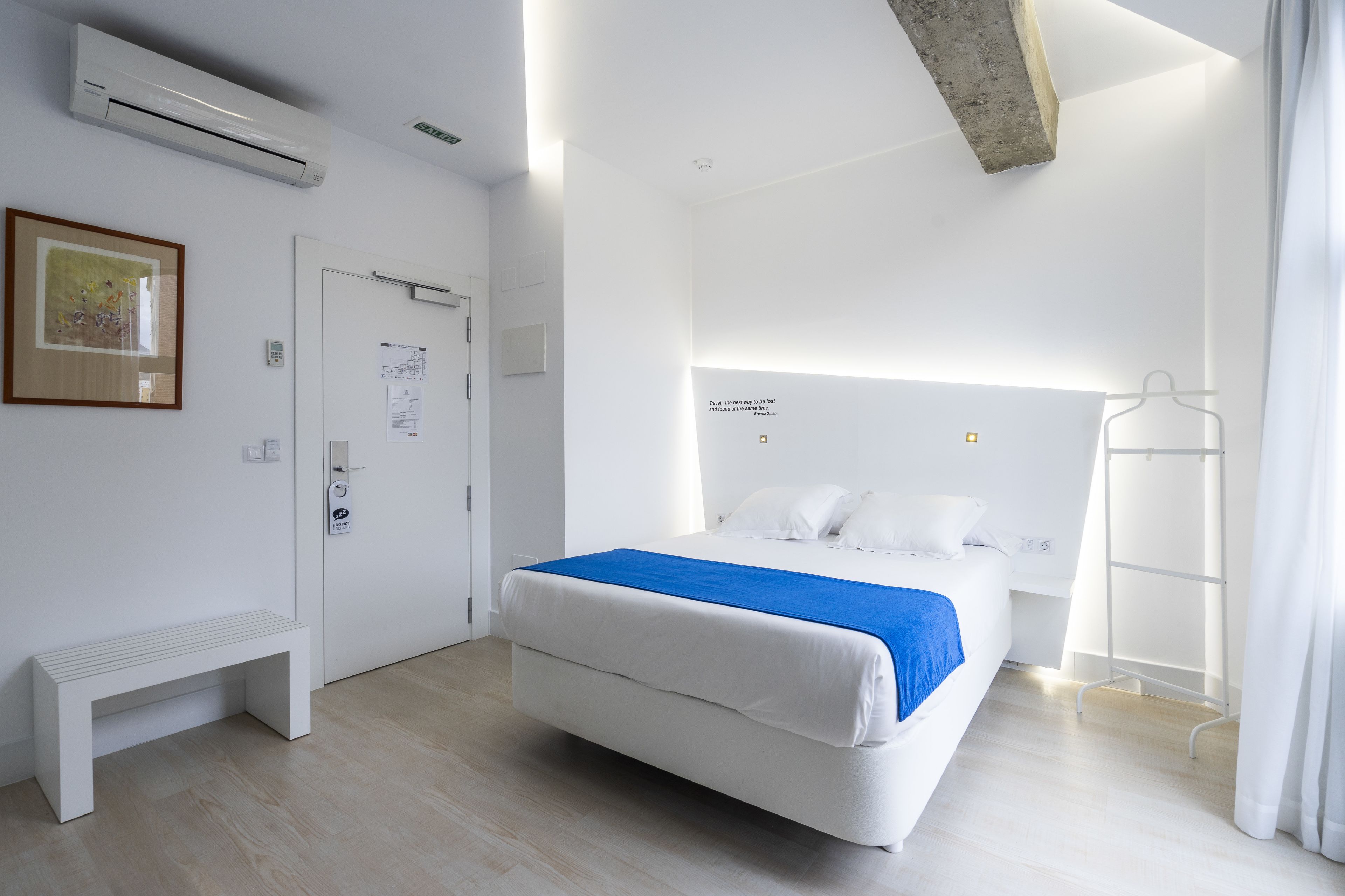Moderni hotellihuone, jossa on sänky, penkki, ilmastointilaite ja naulakko.