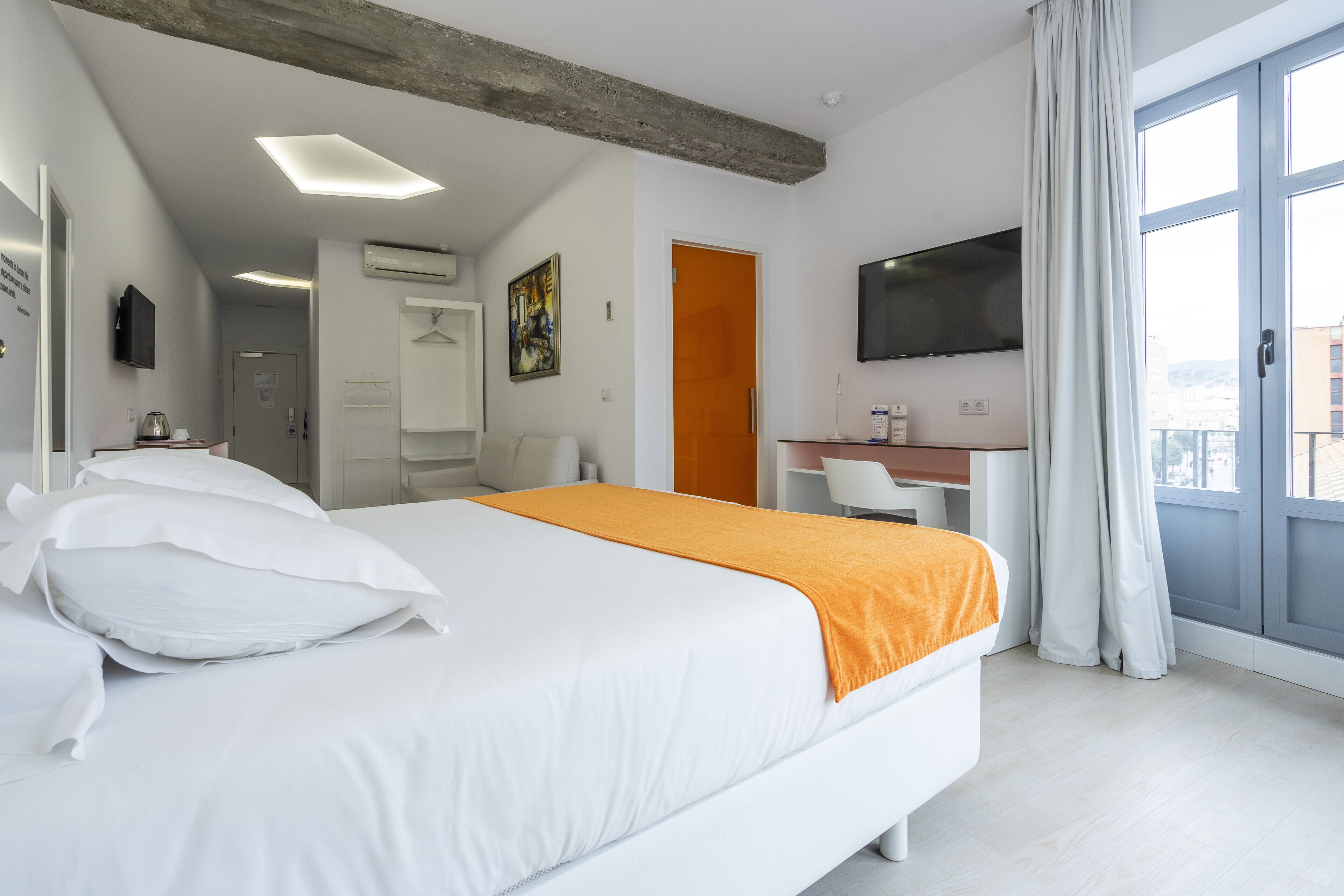 Une chambre d’hôtel avec un grand lit, un plaid orange, un bureau avec une chaise, une télévision et un balcon avec des rideaux.