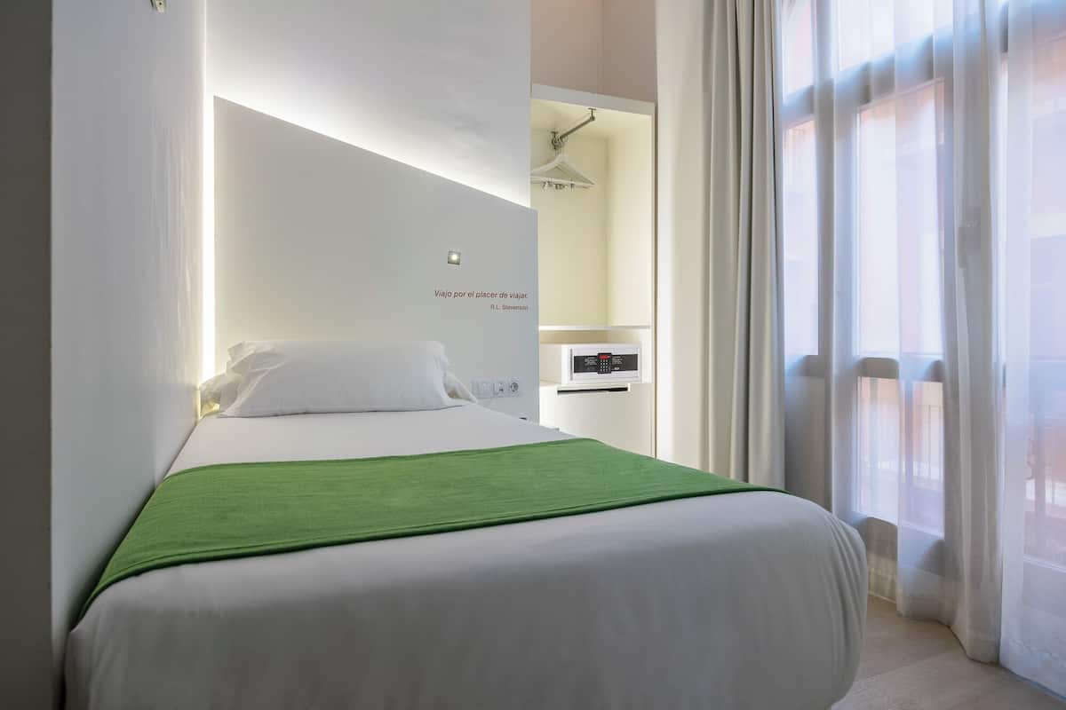 A modern hotel room with a neatly made bed, a green throw blanket, and a wall-mounted control panel.