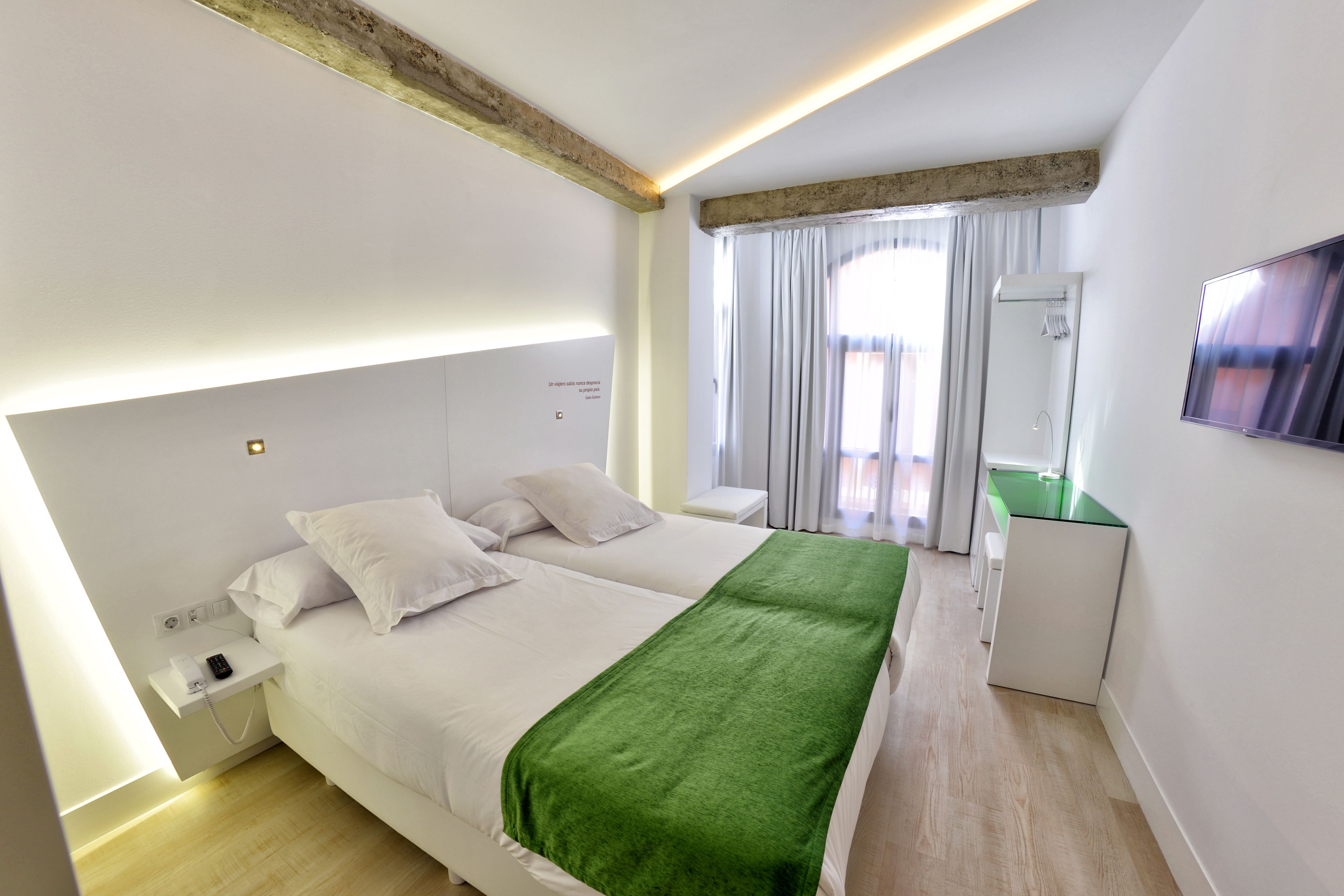 A modern hotel room with a large bed, a green throw blanket, a bedside table with a phone, a built-in TV, and a window with curtains.