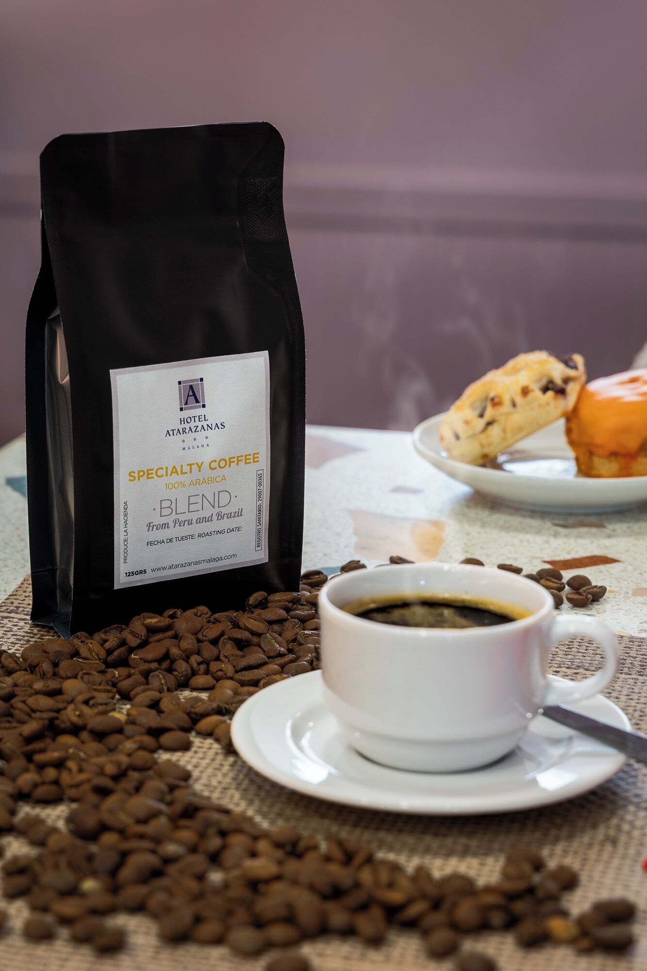 A bag of specialty coffee, a cup of coffee, and a plate with pastries on a table.