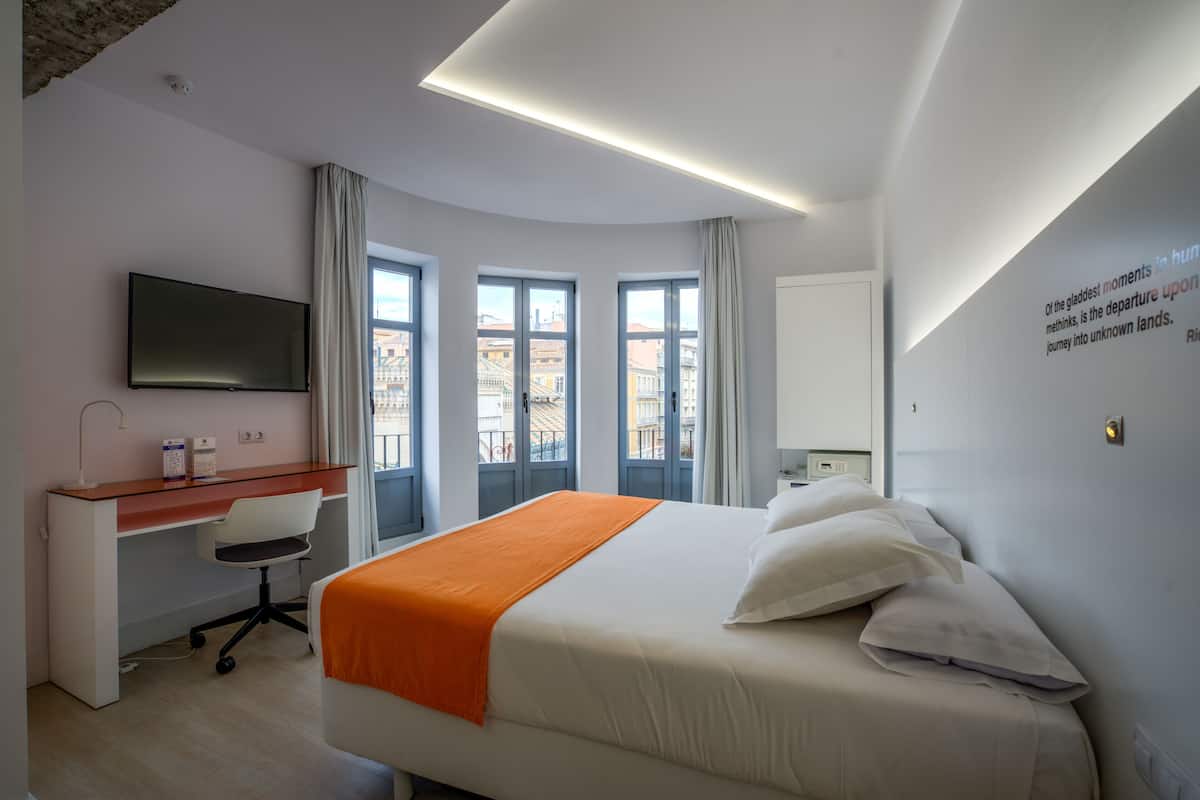 A hotel room with a bed, a desk, a chair, a TV, and large windows.