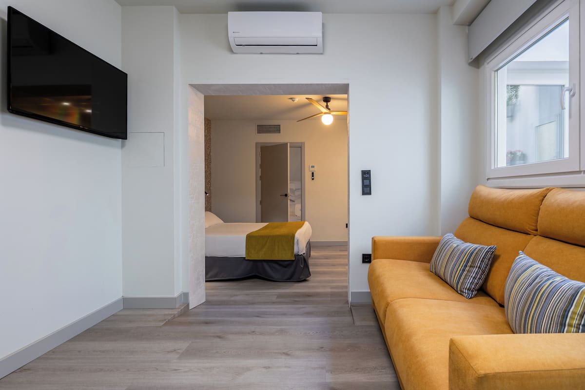 A modern living room with a sofa, a flat-screen TV, and a bed with a yellow blanket.