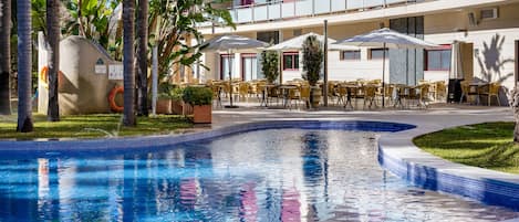 A pool area with a clear blue pool, surrounded by yellow chairs and umbrellas, and a building with balconies in the background.