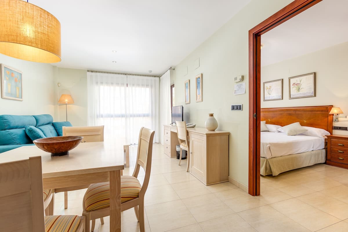 A hotel room with a bed, a desk, a chair, a dining table, and a sofa.