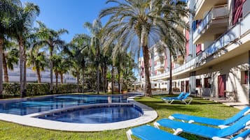 Un espace piscine avec des chaises longues bleues, des palmiers et un bâtiment doté de balcons.