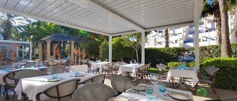 An outdoor dining area with tables set for two, covered by a white canopy, surrounded by chairs and palm trees.