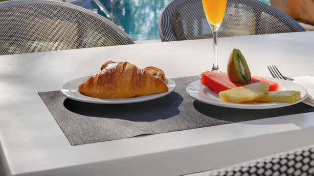 Una mesa con un croissant, una rebanada de sandía, kiwi y un vaso de jugo de naranja, con vista a una piscina, palmeras y un edificio al fondo.