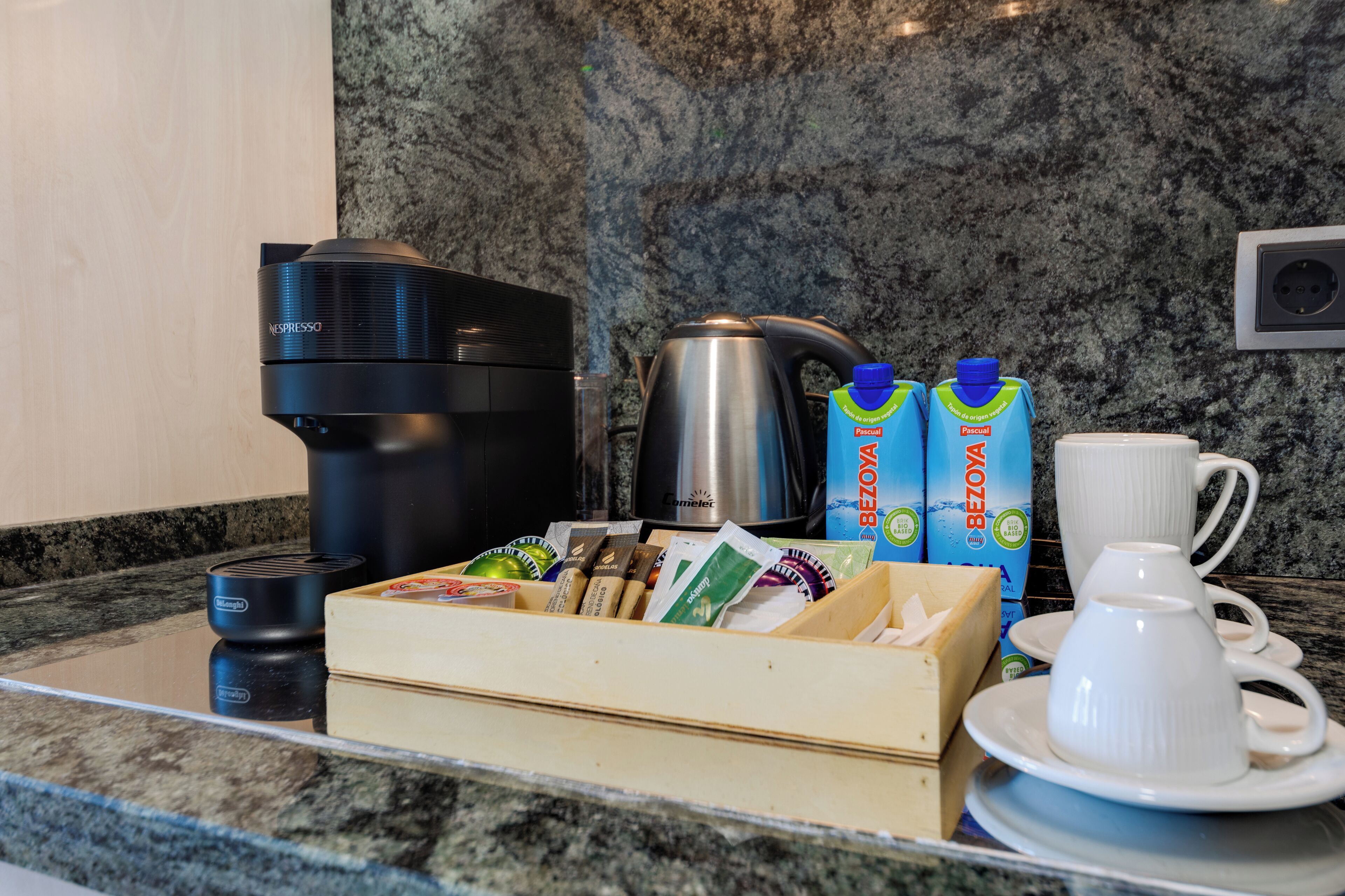 Una encimera de cocina con una cafetera, un hervidor, un cartón de leche, una caja de bolsitas de té y una taza y platillo blancos.