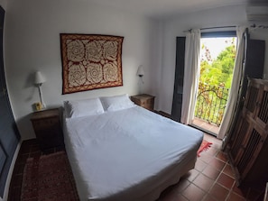 A bedroom with a large bed, a nightstand, a wall-mounted lamp, and a view of greenery through an open door. - Hotel Cerro de Hijar (Tolox)
