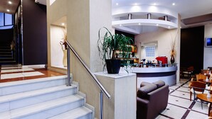 A hotel lobby with a staircase, reception desk, seating area, and a large potted plant. - The Athens Mirabello (Athens)