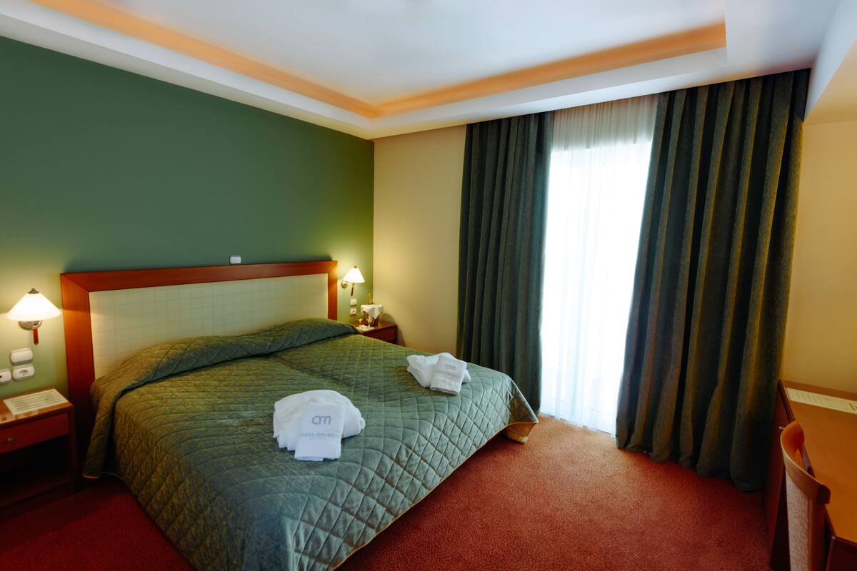 Una habitación de hotel con cama, mesita de noche, lámpara y ventana con cortinas.