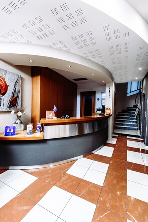 A hotel reception area with a curved counter, a staircase, and a tiled floor. - The Athens Mirabello (Athens)