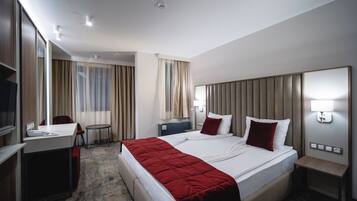 A hotel room with a large bed, a desk, a chair, a small table, a TV, and a mirror.