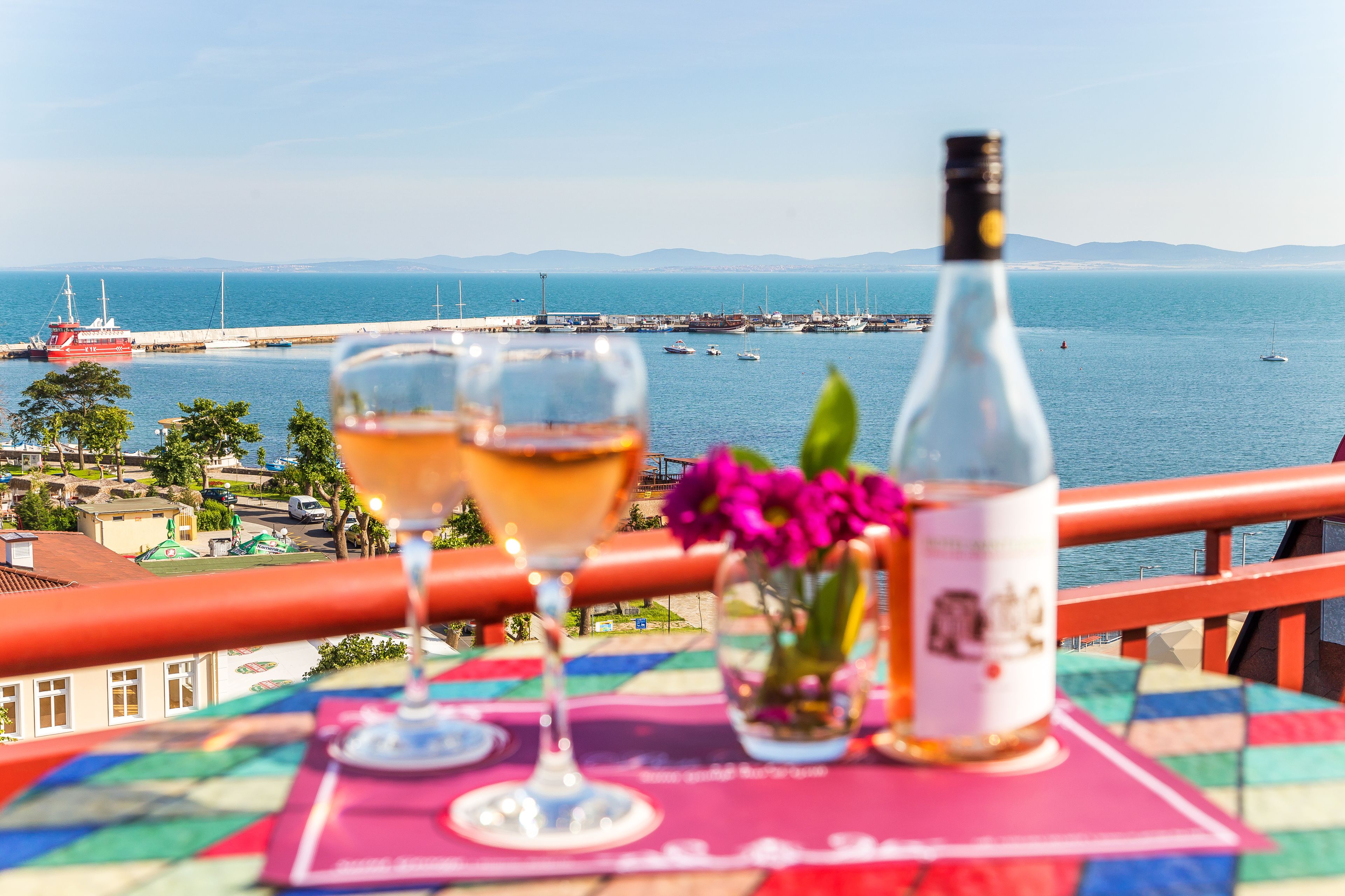 Una mesa con una botella y dos vasos, con vista a un puerto con barcos y una costa lejana.