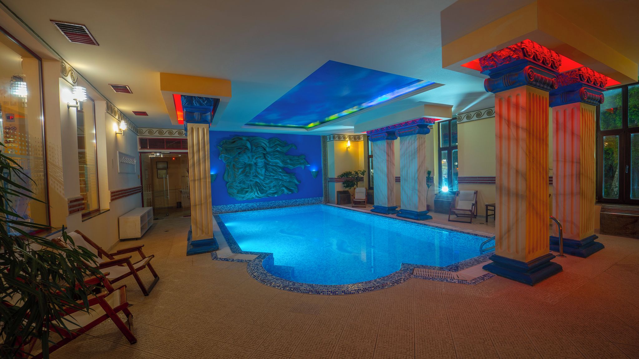 Zwembad met een blauwe mozaïekrand, omringd door kolommen met rode verlichting, en een groot muurdecor.