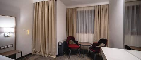 A hotel room with a bed, a desk, two chairs, a heater, and a window with curtains.